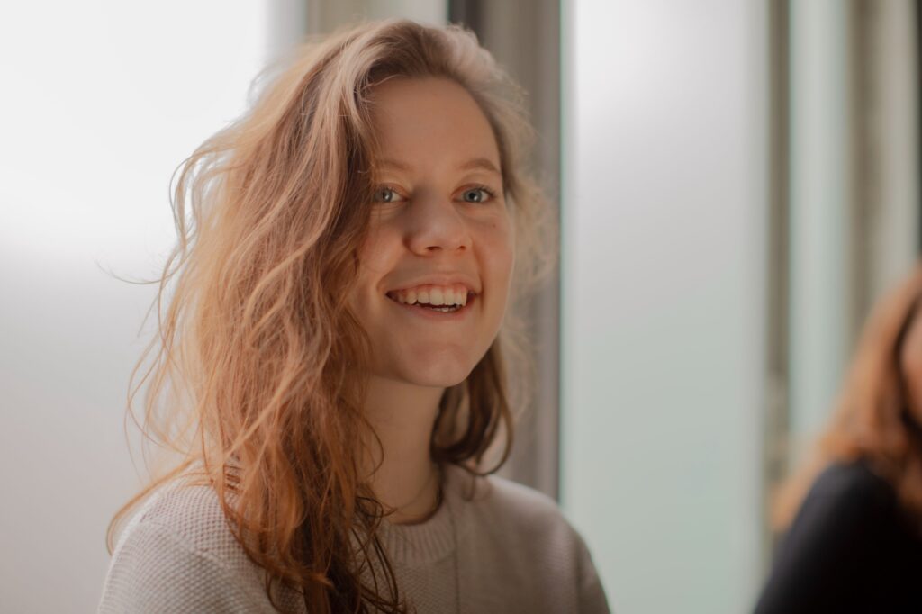 Isis Jonker glimlachend bij een raam, een social media specialist en content creator uit Den Haag, bekend om storytelling en creatieve projecten.